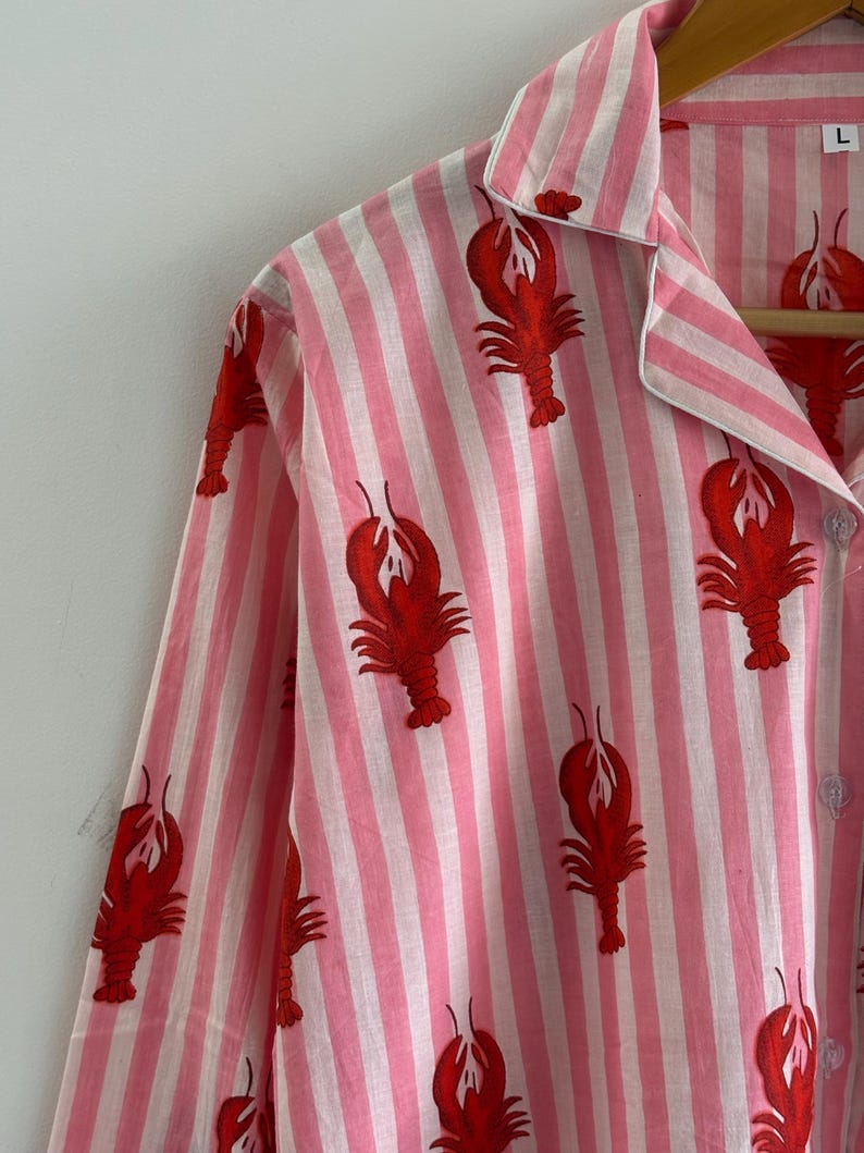 Puede incluir: Camiseta de pijama a rayas rosas y blancas con un estampado de langosta roja. La camiseta de manga larga tiene cuello y se abrocha por delante. El pijama est&aacute; colgado de una percha de madera.