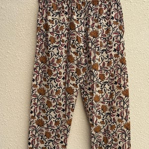 May include: Cream-colored pajama pants with a floral pattern in shades of brown, red, and black. The pants are hanging on a wooden hanger against a neutral wall. The floral design includes stylized flowers and leaves.