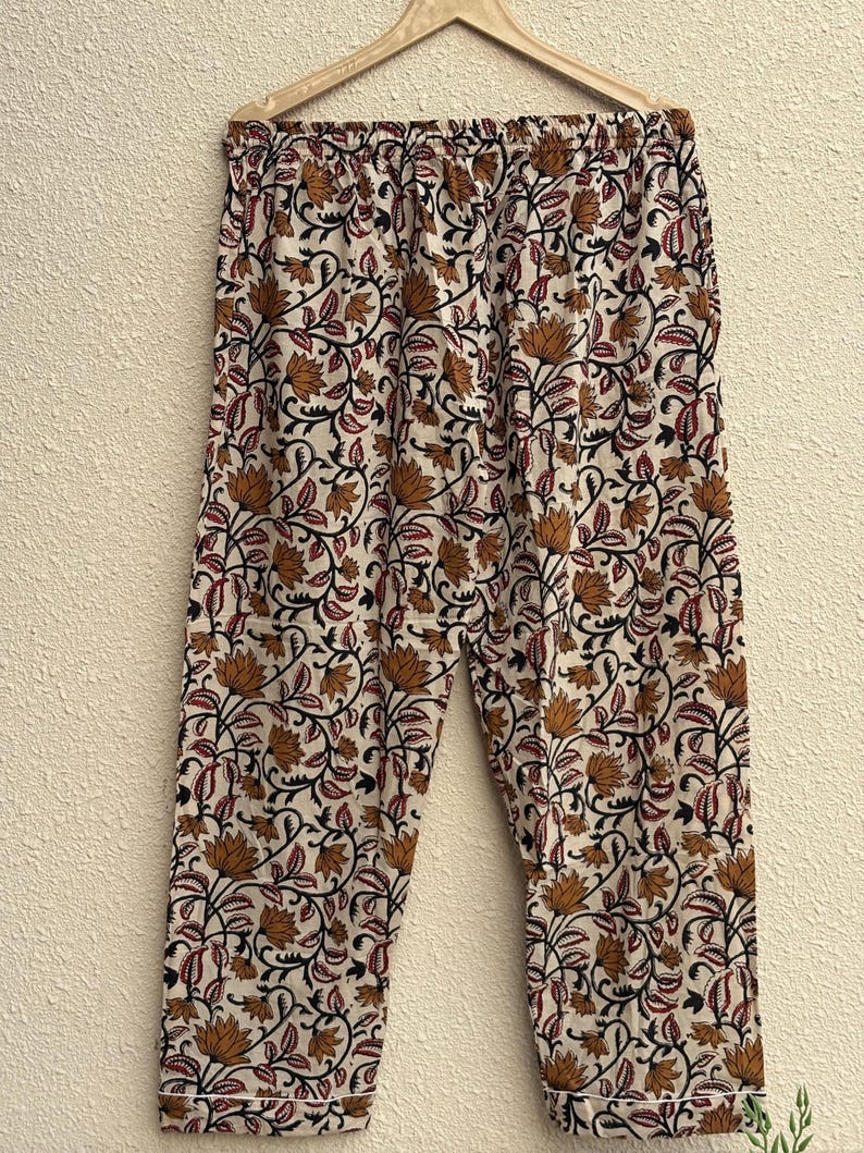 May include: Cream-colored pajama pants with a floral pattern in shades of brown, red, and black. The pants are hanging on a wooden hanger against a neutral wall. The floral design includes stylized flowers and leaves.