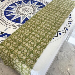 May include: A green and white floral patterned bed runner is draped over a bed. The bedspread underneath features a large blue and white geometric design. The runner's pattern is a repeating floral motif in shades of green and white.