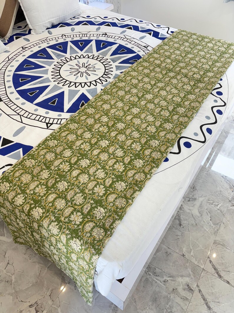 May include: A green and white floral patterned bed runner is draped over a bed. The bedspread underneath features a large blue and white geometric design. The runner's pattern is a repeating floral motif in shades of green and white.