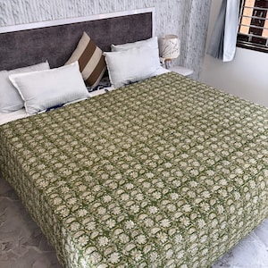 May include: A bed covered with a green floral patterned quilt. The quilt features a repeating white and beige floral design. White pillows and a brown and white striped pillow are arranged on the bed. A gray headboard and a white nightstand are also visible.