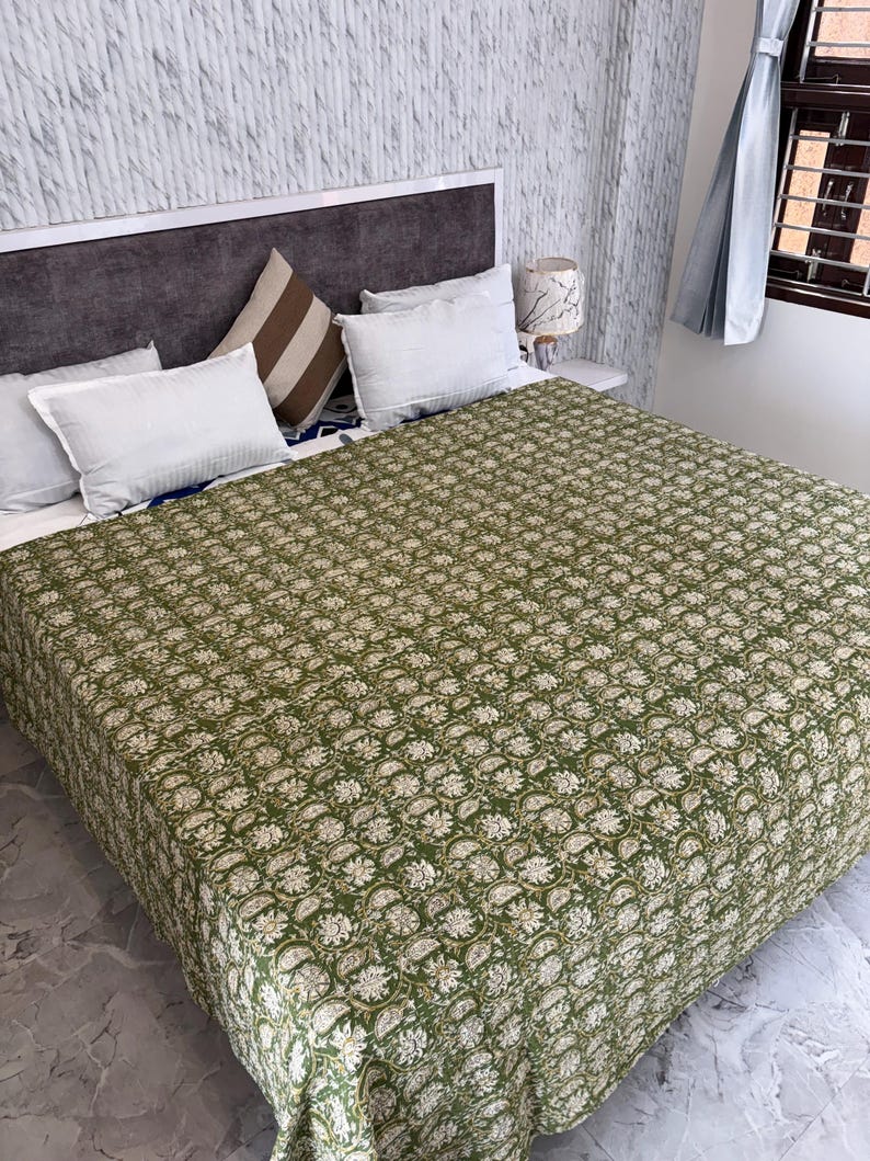 May include: A bed covered with a green floral patterned quilt. The quilt features a repeating white and beige floral design. White pillows and a brown and white striped pillow are arranged on the bed. A gray headboard and a white nightstand are also visible.