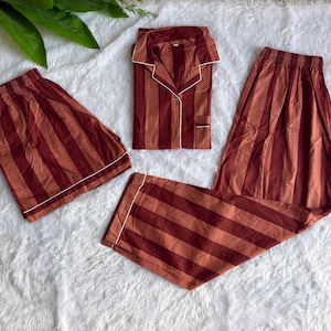 May include: A pajama set consisting of a long-sleeve button-down shirt, a pair of long pants, and a pair of shorts. The garments feature a vertical striped pattern in shades of dark red and light brown with white piping along the edges.