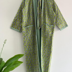 Puede incluir: Una bata larga y abierta con un estampado floral. La bata es principalmente verde lima con un patrón repetido de pequeñas flores moradas y blancas. El interior y el ribete son de color verde claro liso. La bata tiene dos bolsillos y un cinturón a juego.