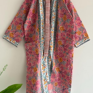 May include: A pink floral patterned robe with a white and teal border. The robe has a relaxed fit with three-quarter sleeves and two pockets. The floral pattern includes yellow and pink flowers.