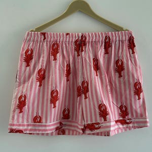Puede incluir: Shorts a rayas rosas y blancas con un patr&oacute;n repetido de langostas rojas. Los shorts tienen una cintura el&aacute;stica y un ribete blanco en el dobladillo inferior. Colgados de una percha de madera.