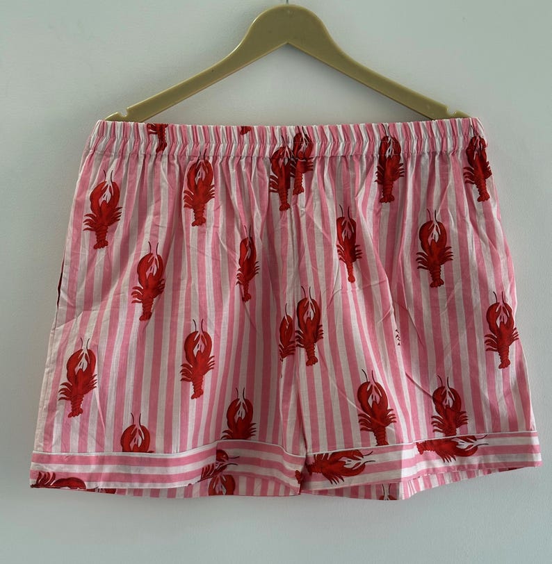 Puede incluir: Shorts a rayas rosas y blancas con un patr&oacute;n repetido de langostas rojas. Los shorts tienen una cintura el&aacute;stica y un ribete blanco en el dobladillo inferior. Colgados de una percha de madera.