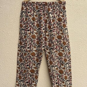 May include: Cream-colored pajama pants with a repeating floral pattern in shades of brown, black, and red. The pants are hanging on a wooden hanger. The floral design includes stylized lotus flowers and vines.