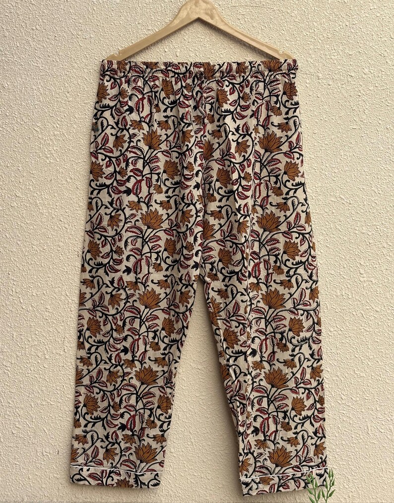 May include: Cream-colored pajama pants with a repeating floral pattern in shades of brown, black, and red. The pants are hanging on a wooden hanger. The floral design includes stylized lotus flowers and vines.
