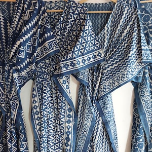 May include: Several indigo blue and white patterned robes hang on wooden hangers. The robes feature various geometric and floral designs. The fabric appears to be lightweight, with a flowing drape. The robes are displayed against a plain white background.