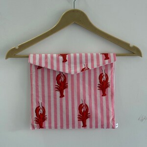 Puede incluir: Una bolsa de tela a rayas rosas y blancas con motivos de langosta roja, colgada de una percha de madera. La bolsa tiene un cierre de solapa. El dise&ntilde;o es de tem&aacute;tica n&aacute;utica y el fondo es una pared blanca.