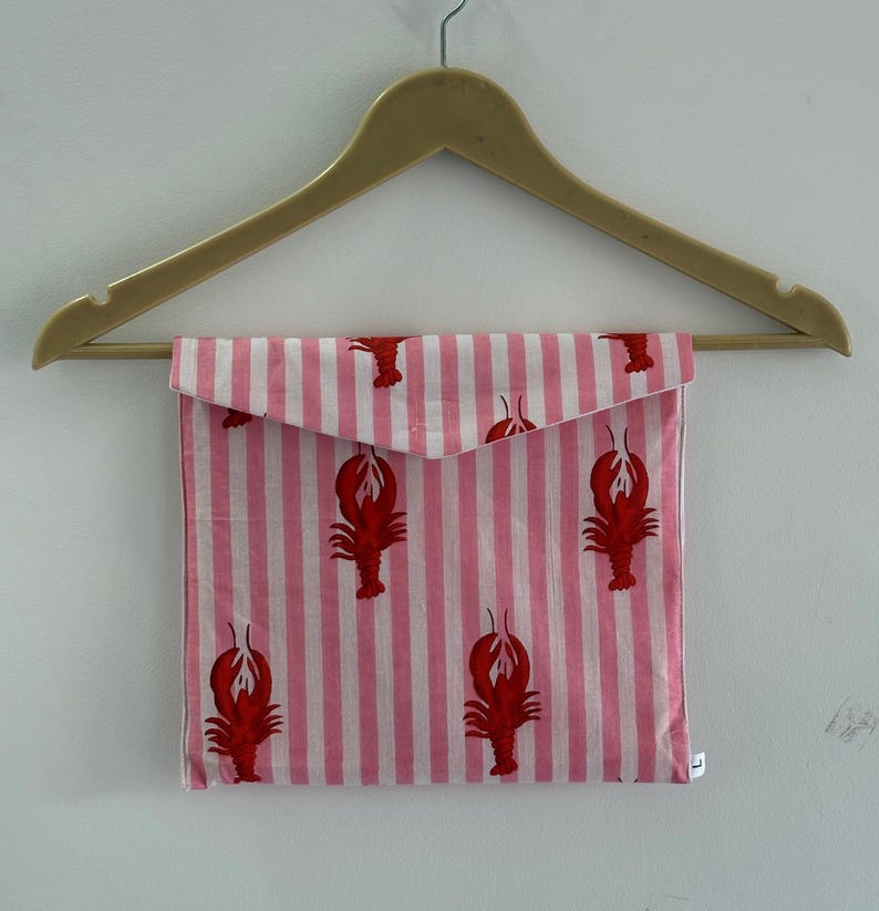 Puede incluir: Una bolsa de tela a rayas rosas y blancas con motivos de langosta roja, colgada de una percha de madera. La bolsa tiene un cierre de solapa. El dise&ntilde;o es de tem&aacute;tica n&aacute;utica y el fondo es una pared blanca.