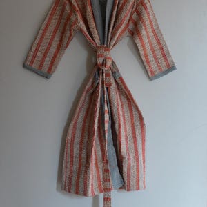 May include: A striped robe with a chevron pattern in orange and beige, with gray trim and a matching belt. The robe is hanging on a wooden hanger against a light gray wall.