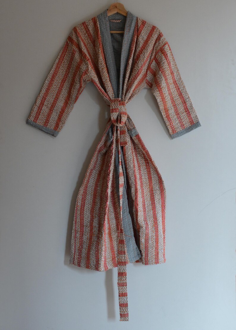 May include: A striped robe with a chevron pattern in orange and beige, with gray trim and a matching belt. The robe is hanging on a wooden hanger against a light gray wall.