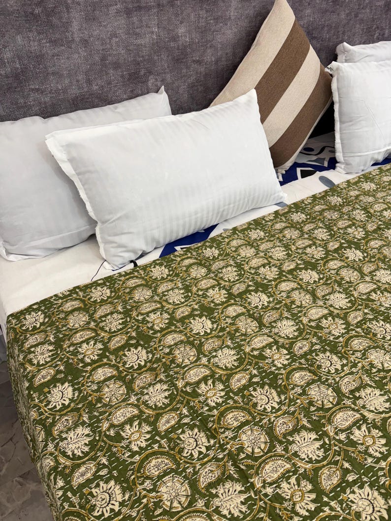 High-angle view of a sage green floral kantha quilt with white and gold block print patterns, spread neatly on a large bed with white and brown striped pillows against a textured grey wall.