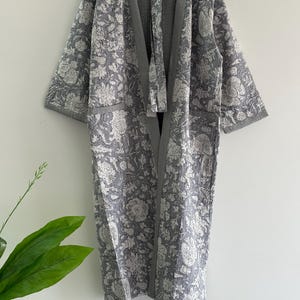 May include: A gray and white floral print kimono robe. The robe features a long, open design with a tie closure and pockets. The floral pattern is a mix of large and small blooms. The robe is made of a soft, quilted fabric.