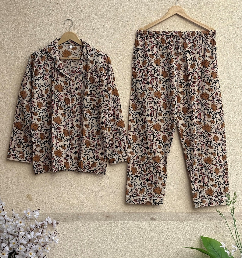 Women's 3-piece loungewear set featuring a long-sleeve button-down shirt and matching trousers. The design is a traditional Indian floral vine block print on a beige base. Displayed on wooden hangers against a textured wall.