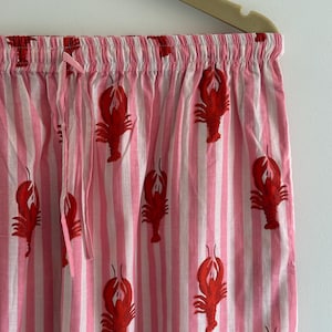 Puede incluir: Pantalones de pijama a rayas rosas y blancas con un estampado repetido de langostas rojas. Los pantalones tienen cintura con cord&oacute;n y est&aacute;n colgados de una percha beige. La tela es ligera y parece ser de algod&oacute;n.