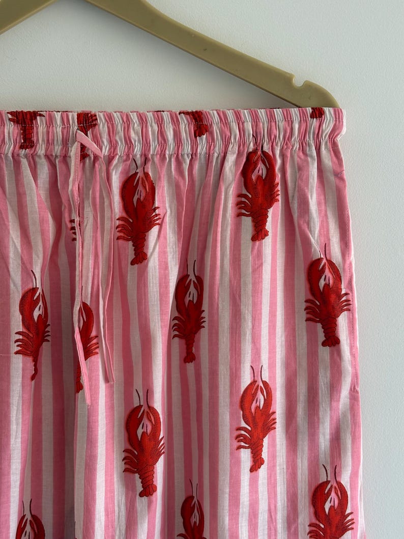 Puede incluir: Pantalones de pijama a rayas rosas y blancas con un estampado repetido de langostas rojas. Los pantalones tienen cintura con cord&oacute;n y est&aacute;n colgados de una percha beige. La tela es ligera y parece ser de algod&oacute;n.