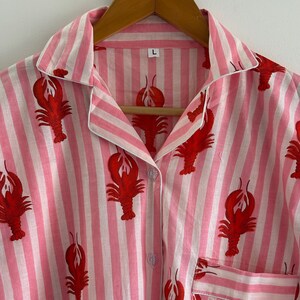 Puede incluir: Camiseta de pijama a rayas rosas y blancas con un estampado de langosta roja. La camiseta tiene cuello, cierre de botones y bolsillo en el pecho. La talla "L" es visible en la etiqueta. La prenda est&aacute; colgada en una percha de madera.