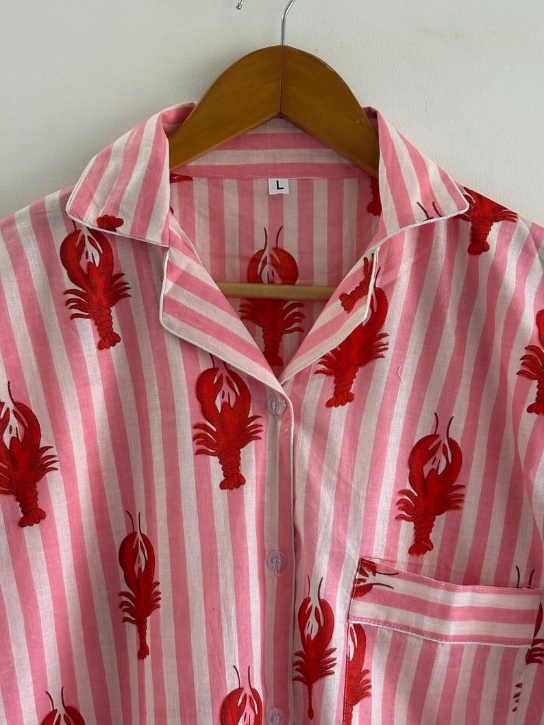 Puede incluir: Camiseta de pijama a rayas rosas y blancas con un estampado de langosta roja. La camiseta tiene cuello, cierre de botones y bolsillo en el pecho. La talla "L" es visible en la etiqueta. La prenda est&aacute; colgada en una percha de madera.