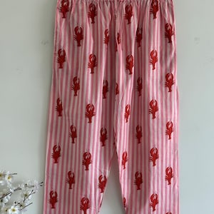 Puede incluir: Pantalones de pijama a rayas rosas y blancas con un estampado repetido de langostas rojas. Los pantalones cuelgan de una percha de madera sobre un fondo blanco. Los pantalones de pijama tienen cintura con cord&oacute;n.