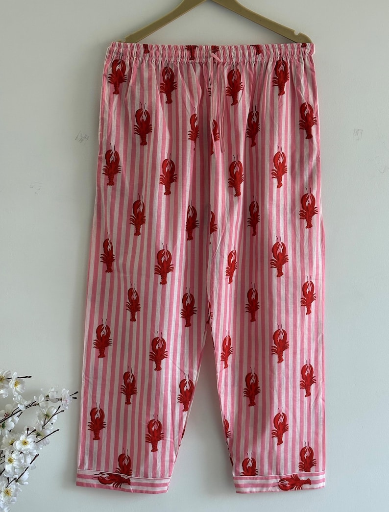 Puede incluir: Pantalones de pijama a rayas rosas y blancas con un estampado repetido de langostas rojas. Los pantalones cuelgan de una percha de madera sobre un fondo blanco. Los pantalones de pijama tienen cintura con cord&oacute;n.