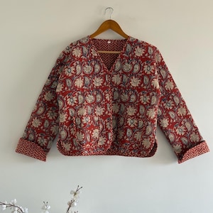 Op de afbeelding: Een rode gewatteerde jas met een bloemenpatroon in crème, blauw en groen. De jas heeft lange mouwen met omgeslagen manchetten en een V-hals. Hij hangt aan een houten hanger tegen een witte achtergrond.