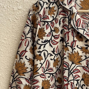 May include: Cream-colored button-down shirt with a floral pattern. The design features brown and red flowers with black stems and leaves. The shirt has a white collar and cuffs, and is hanging on a wooden hanger.