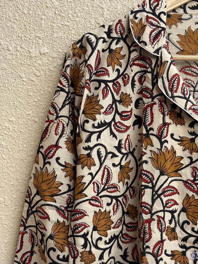 May include: Cream-colored button-down shirt with a floral pattern. The design features brown and red flowers with black stems and leaves. The shirt has a white collar and cuffs, and is hanging on a wooden hanger.