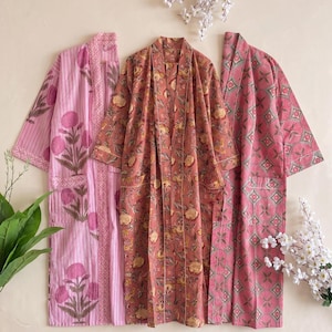 Könnte beinhalten: Drei langärmelige Morgenmäntel mit unterschiedlichen Mustern: ein hellrosa Modell mit vertikalen Streifen und Blumenmotiven, ein terrakottafarbenes Modell mit gelbem Blumendruck und ein rosa Modell mit geometrischem Rauten- und Blumenmuster.