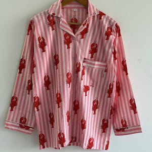 Puede incluir: Camisa de pijama a rayas rosas y blancas con un estampado de langostas rojas. La camisa de manga larga tiene cuello y bolsillo en el pecho. La prenda est&aacute; colgada de una percha de madera y est&aacute; hecha de tela ligera.