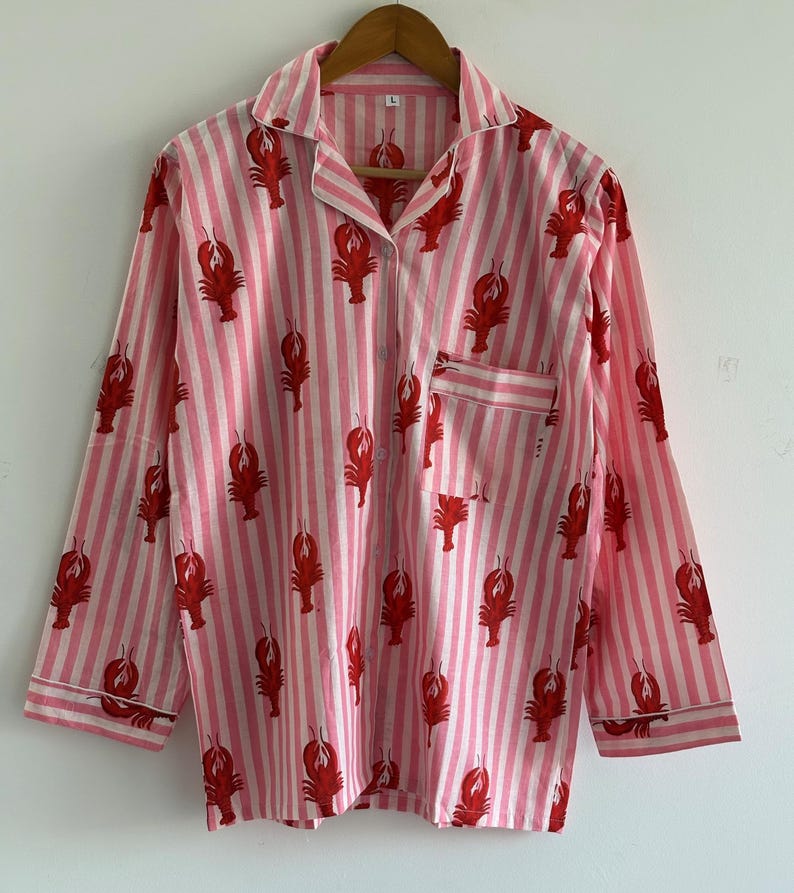 Puede incluir: Camisa de pijama a rayas rosas y blancas con un estampado de langostas rojas. La camisa de manga larga tiene cuello y bolsillo en el pecho. La prenda est&aacute; colgada de una percha de madera y est&aacute; hecha de tela ligera.