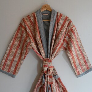 May include: A handmade robe with a chevron pattern in orange, beige, and gray. The robe is hanging on a wooden hanger. The robe has a tie closure and gray trim.