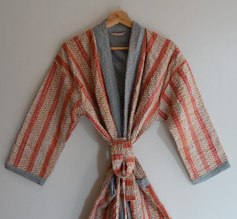 May include: A handmade robe with a chevron pattern in orange, beige, and gray. The robe is hanging on a wooden hanger. The robe has a tie closure and gray trim.