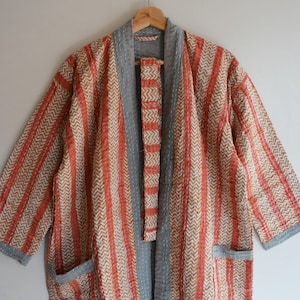 May include: A patterned, open-front robe with a gray collar and trim, hanging on a wooden hanger. The robe features a combination of vertical stripes and chevron patterns in shades of orange, beige, and gray. It has two front pockets.