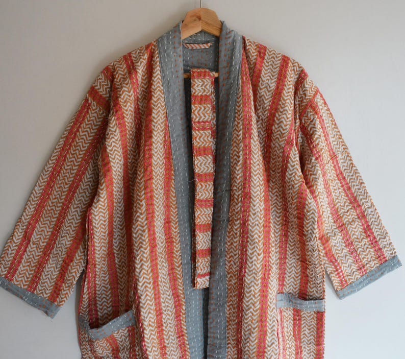 May include: A patterned, open-front robe with a gray collar and trim, hanging on a wooden hanger. The robe features a combination of vertical stripes and chevron patterns in shades of orange, beige, and gray. It has two front pockets.