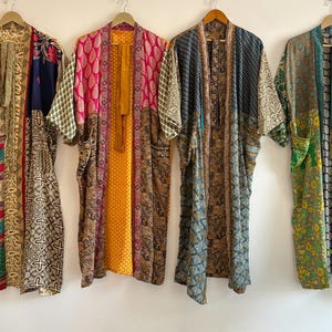 May include: Four colorful, patterned kimono robes hang against a white wall. The robes feature a variety of designs, including floral, geometric, and patchwork patterns. Each robe has a unique color scheme, with hues of blue, pink, yellow, and green.