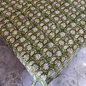 May include: A green tablecloth with a repeating floral pattern in white and gold. The fabric is draped over a table, showcasing the intricate design and texture of the cloth. The floral pattern includes paisley and circular elements.