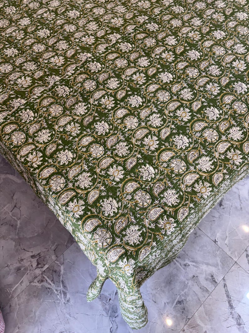 May include: A green tablecloth with a repeating floral pattern in white and gold. The fabric is draped over a table, showcasing the intricate design and texture of the cloth. The floral pattern includes paisley and circular elements.