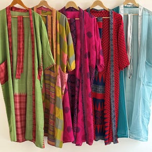 May include: A collection of colorful, patterned kimono robes hanging on wooden hangers. The robes feature a variety of designs, including floral, geometric, and patchwork patterns. Colors include green, pink, red, blue, and yellow. The robes are made of lightweight fabric.