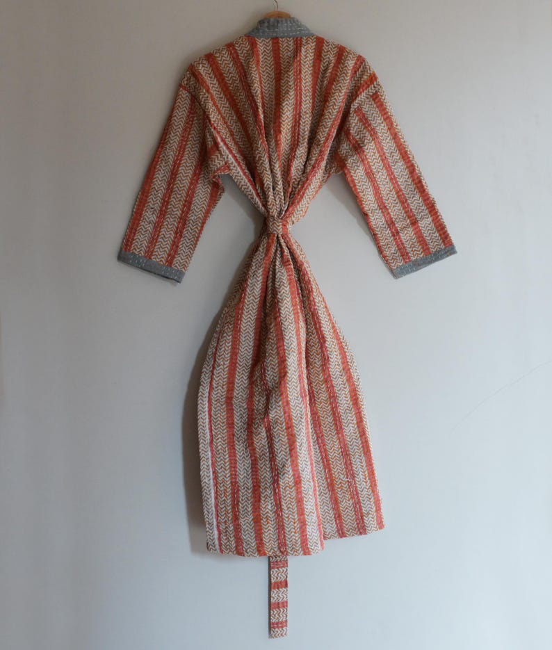 May include: A patterned robe with vertical stripes in shades of orange, beige, and gray. The robe is tied at the waist and has long sleeves with gray cuffs. The robe is hanging on a wooden hanger.