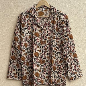 May include: Cream-colored pajama set with a floral pattern in shades of brown, black, and red. The long-sleeved top has a collared neckline, a front pocket, and white piping along the edges. The set is hanging on a wooden hanger.
