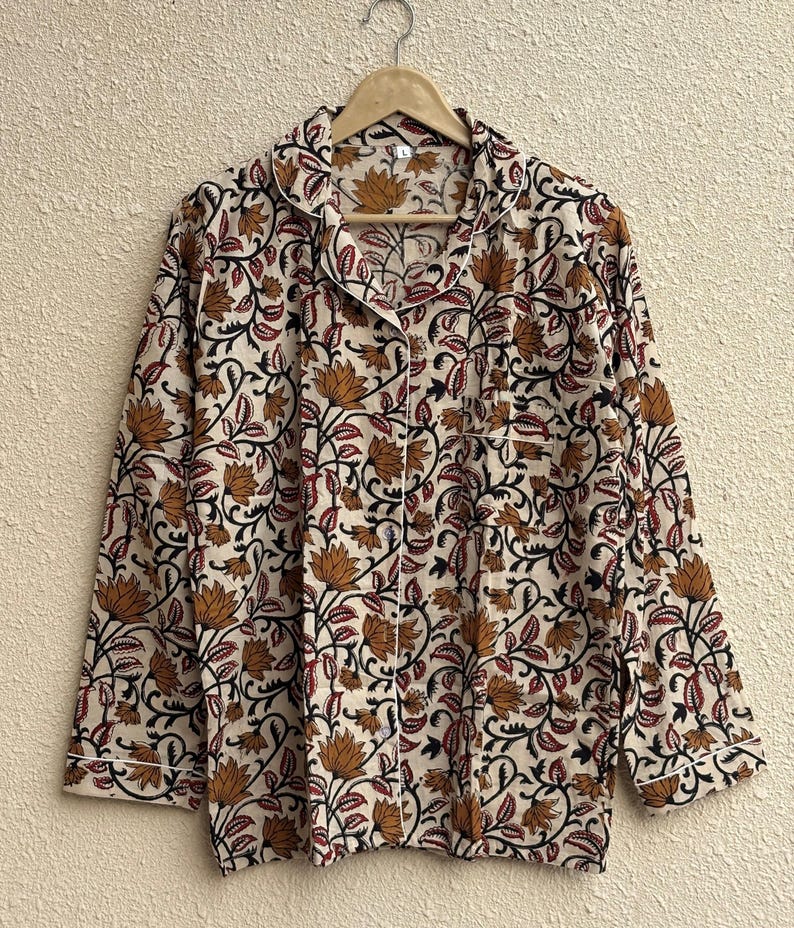 May include: Cream-colored pajama set with a floral pattern in shades of brown, black, and red. The long-sleeved top has a collared neckline, a front pocket, and white piping along the edges. The set is hanging on a wooden hanger.