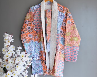 Puede incluir: Bata tipo kimono patchwork con un estampado floral en tonos naranja, azul y rosa. La bata tiene un ribete blanco y está colgada de una percha de madera. La bata está hecha de una tela ligera.