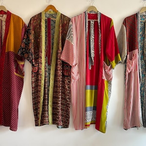 Peut inclure: Quatre kimonos patchwork colorés sont suspendus à des cintres en bois contre un mur blanc. Les kimonos présentent divers motifs, notamment des rayures et des motifs floraux. Les couleurs incluent le rouge, le jaune, le marron et le vert.
