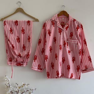 Two-piece women's pajama set featuring red lobster print on pink and white vertical stripes. Includes a long-sleeve button-down shirt and full-length trousers hanging on wooden hangers.