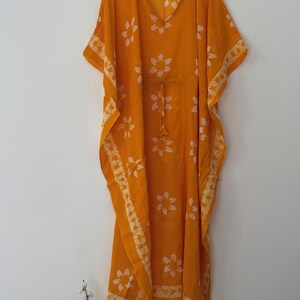 Op de afbeelding: Een oranje kaftan jurk met witte bloempatronen. De jurk heeft een V-hals, korte mouwen en een taille met trekkoord. Het bloemmotief wordt herhaald op de mouwen en zoom. De kaftan hangt aan een houten hanger.