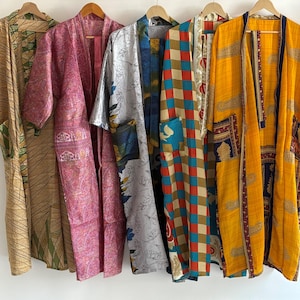 Könnte beinhalten: Eine Sammlung von fünf farbenfrohen, gemusterten Kimonos, die an Holzbügeln hängen. Die Kimonos weisen verschiedene Designs auf, darunter florale, geometrische und abstrakte Muster in Gelb-, Rosa-, Blau- und Brauntönen. Jeder Kimono hat einen einzigartigen Stil und eine einzigartige Farbpalette.